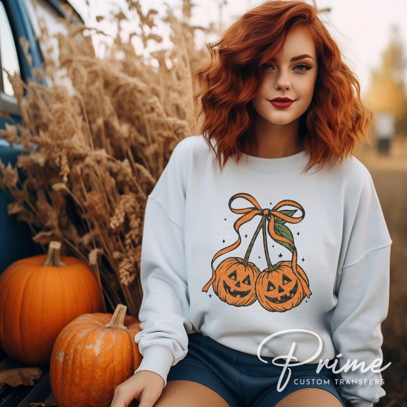 May include: A light blue sweatshirt featuring a Halloween-themed design of two jack-o'-lanterns tied together with an orange bow. Two orange pumpkins are in the foreground. The sweatshirt is part of a fall-themed collection.
