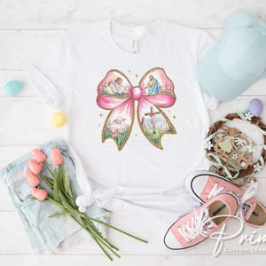 K&ouml;nnte beinhalten: Ein wei&szlig;es T-Shirt mit einem rosafarbenen Schleifendesign, das eine religi&ouml;se Szene mit einem Lamm, einem Kreuz und einer Taube zeigt. Die Schleife ist von einem gr&uuml;nen floralen Rand umgeben.