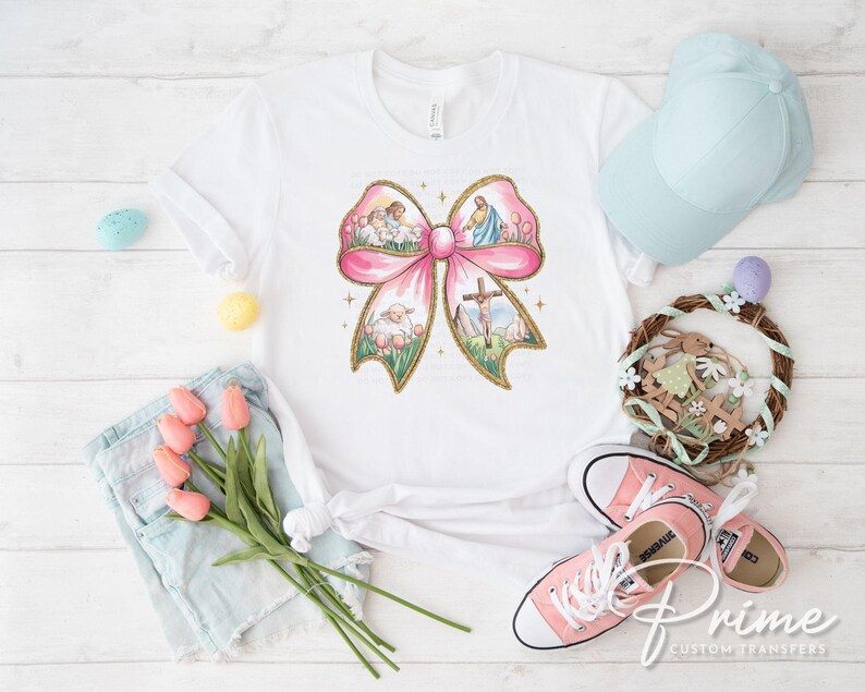 K&ouml;nnte beinhalten: Ein wei&szlig;es T-Shirt mit einem rosafarbenen Schleifendesign, das eine religi&ouml;se Szene mit einem Lamm, einem Kreuz und einer Taube zeigt. Die Schleife ist von einem gr&uuml;nen floralen Rand umgeben.