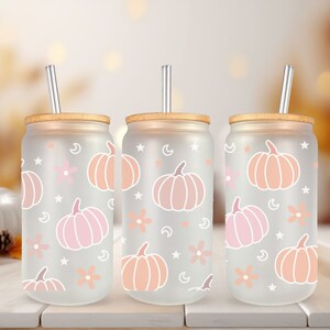 K&ouml;nnte beinhalten: Drei gefrostete Glasbecher mit Bambusdeckeln und klaren Strohhalmen. Jeder Becher zeigt ein wei&szlig;es K&uuml;rbis- und Blumenmuster auf einem hellrosa Hintergrund.