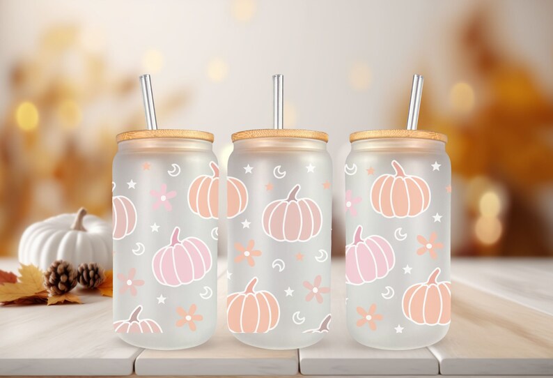 K&ouml;nnte beinhalten: Drei gefrostete Glasbecher mit Bambusdeckeln und klaren Strohhalmen. Jeder Becher zeigt ein wei&szlig;es K&uuml;rbis- und Blumenmuster auf einem hellrosa Hintergrund.