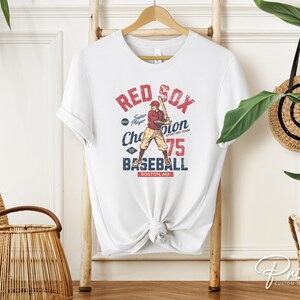 Może przedstawiać: Biała koszulka z grafiką w stylu vintage przedstawiającą baseballistę. Grafika zawiera napis "Red Sox" i "Baseball" oraz tekst "Super Player Champion Sporting Team 75 Boston, MA".