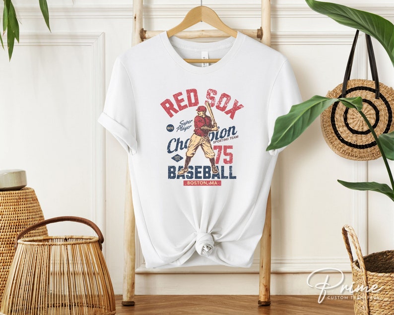 Może przedstawiać: Biała koszulka z grafiką w stylu vintage przedstawiającą baseballistę. Grafika zawiera napis "Red Sox" i "Baseball" oraz tekst "Super Player Champion Sporting Team 75 Boston, MA".