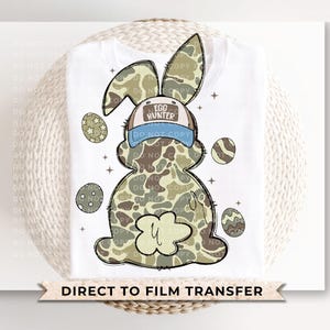 May include: A white t-shirt with a camouflage bunny rabbit wearing a blue and white hat that says "Egg Hunter". The bunny is surrounded by colorful Easter eggs.