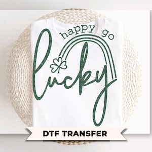 Op de afbeelding: Wit T-shirt met een groen ontwerp met de tekst "happy go lucky" met een klavertje vier en een regenboog. De woorden "DTF TRANSFER" staan onderaan de afbeelding.