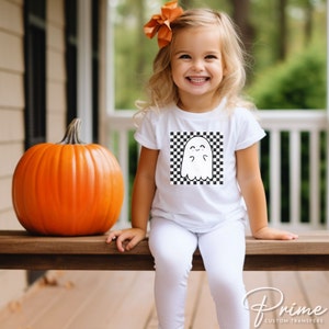 May include: A white t-shirt with a black and white checkered design featuring a cartoon ghost. The ghost has a happy expression and is wearing a bow.
