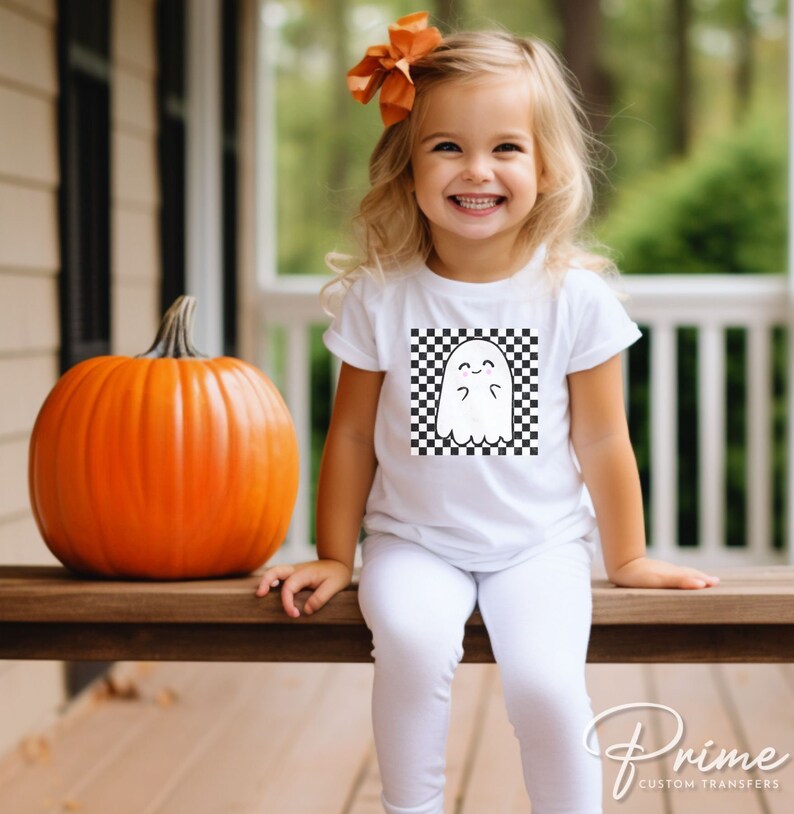May include: A white t-shirt with a black and white checkered design featuring a cartoon ghost. The ghost has a happy expression and is wearing a bow.