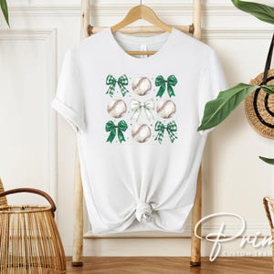 May include: White t-shirt with a green and white baseball and bow design. The design features multiple baseballs and bows in a repeating pattern.
