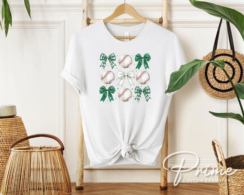 May include: White t-shirt with a green and white baseball and bow design. The design features multiple baseballs and bows in a repeating pattern.