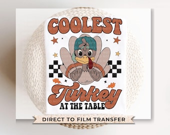 May include: A white iron-on transfer design featuring a cartoon turkey wearing a blue baseball cap and the text "Coolest Turkey at the Table".