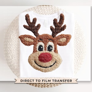 May include: White t-shirt featuring a detailed, textured reindeer face with brown antlers, tan ears, and a red nose. The reindeer has black eyes and a smiling mouth. The text "DIRECT TO FILM TRANSFER" is at the bottom.
