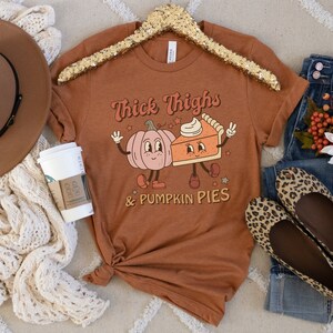 K&ouml;nnte beinhalten: Ein braunes T-Shirt mit dem Text "Thick Thighs & Pumpkin Pies" in Wei&szlig;. Das Shirt zeigt eine Cartoon-K&uuml;rbis und ein St&uuml;ck K&uuml;rbiskuchen mit Gesichtern.