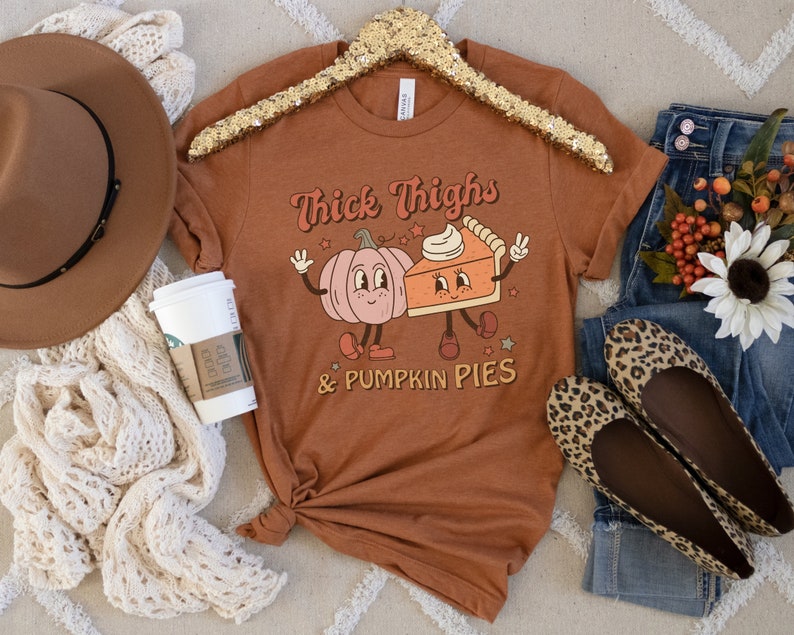 K&ouml;nnte beinhalten: Ein braunes T-Shirt mit dem Text "Thick Thighs & Pumpkin Pies" in Wei&szlig;. Das Shirt zeigt eine Cartoon-K&uuml;rbis und ein St&uuml;ck K&uuml;rbiskuchen mit Gesichtern.