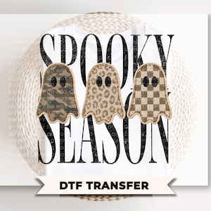 May include: A white t-shirt featuring the black text SPOOKY SEASON and three ghost graphics. The ghosts have distinct patterns: one with camouflage, one with a leopard print, and one with a brown and tan checkerboard pattern, all outlined in tan.