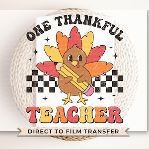 May include: A white iron-on transfer design featuring a cartoon turkey holding a pencil with the text "One Thankful Teacher" in a retro font. The design also includes a checkered pattern and stars.