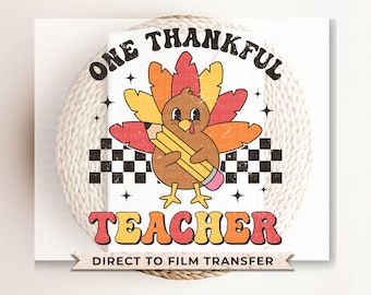 Op de afbeelding: Een wit strijktransferontwerp met een cartoon-kalkoen die een potlood vasthoudt met de tekst "One Thankful Teacher" in een retro-lettertype. Het ontwerp bevat ook een geruit patroon en sterren.
