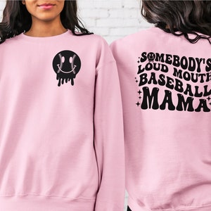 May include: Pink sweatshirt with a black baseball-themed smiley face on the front and the text "Somebody's Loud Mouth Baseball Mama" on the back.
