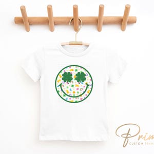 May include: White t-shirt with a St. Patrick's Day design. The design features a smiley face with shamrock eyes and a green outline. The face is filled with rainbows, pots of gold, and other St. Patrick's Day symbols.