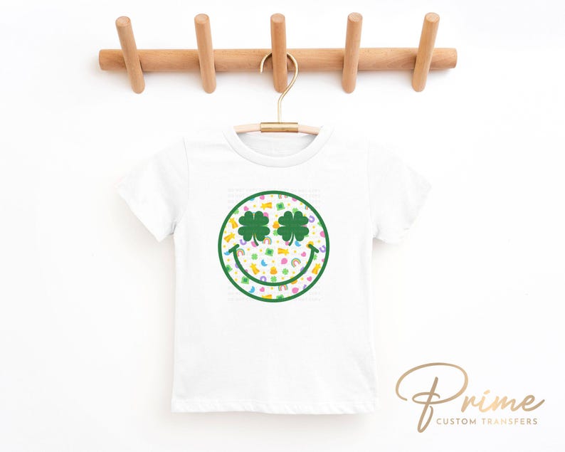 May include: White t-shirt with a St. Patrick's Day design. The design features a smiley face with shamrock eyes and a green outline. The face is filled with rainbows, pots of gold, and other St. Patrick's Day symbols.