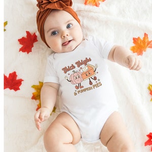 K&ouml;nnte beinhalten: Ein wei&szlig;er Baby-Strampler mit einer niedlichen Grafik von einem K&uuml;rbis und einem K&uuml;rbiskuchen mit dem Text "Thick Thighs & Pumpkin Pies".