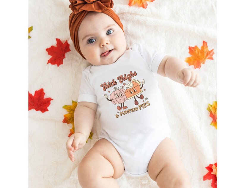 K&ouml;nnte beinhalten: Ein wei&szlig;er Baby-Strampler mit einer niedlichen Grafik von einem K&uuml;rbis und einem K&uuml;rbiskuchen mit dem Text "Thick Thighs & Pumpkin Pies".