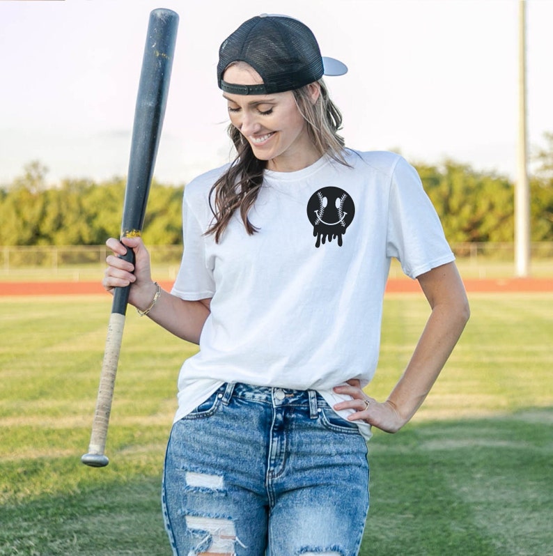 May include: A white t-shirt with a black baseball-shaped smiley face graphic. The person is wearing blue jeans and a black baseball cap.