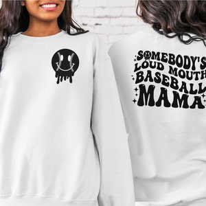 May include: White sweatshirt with a black baseball-themed smiley face on the front and the text "Somebody's Loud Mouth Baseball Mama" on the back.