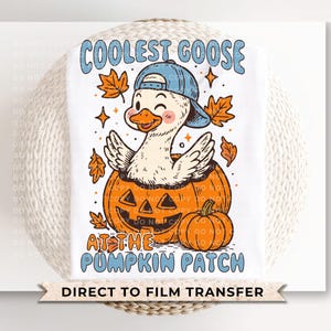 May include: White t-shirt featuring a cartoon goose wearing a blue cap, emerging from an orange jack-o'-lantern. The text reads "COOLEST GOOSE AT THE PUMPKIN PATCH". Autumn leaves and stars surround the goose and pumpkin. A small pumpkin is next to the jack-o'-lantern.