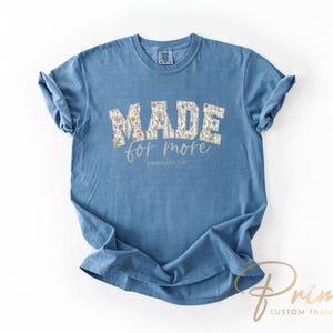 Puede incluir: Una camiseta azul con la frase "MADE for more" en un dise&ntilde;o floral. El texto "EPHESIANS 2:10" est&aacute; debajo. La camiseta tiene las mangas enrolladas y se muestra sobre un fondo blanco.