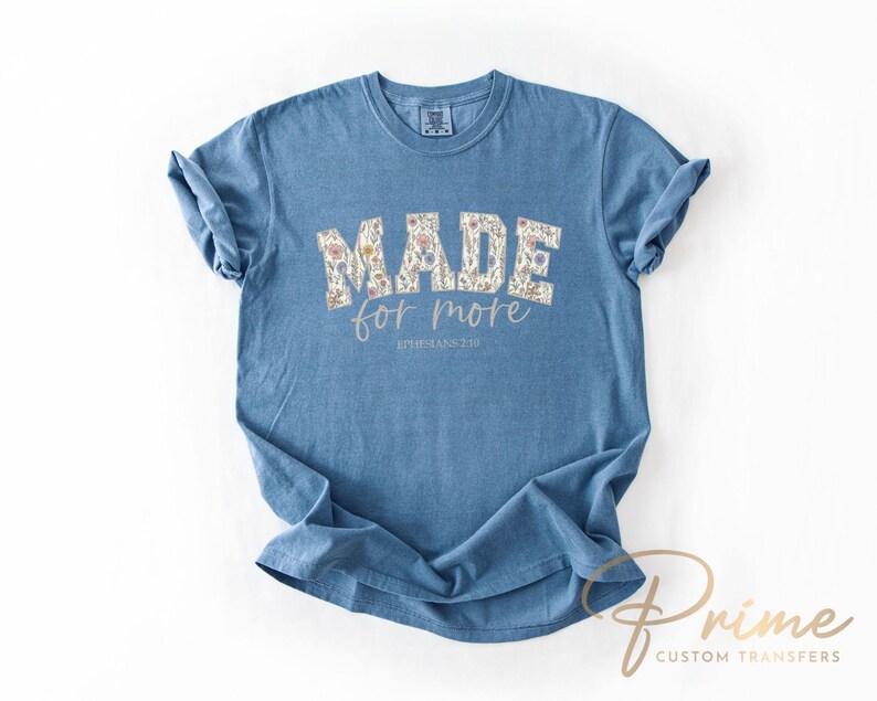 Puede incluir: Una camiseta azul con la frase "MADE for more" en un dise&ntilde;o floral. El texto "EPHESIANS 2:10" est&aacute; debajo. La camiseta tiene las mangas enrolladas y se muestra sobre un fondo blanco.