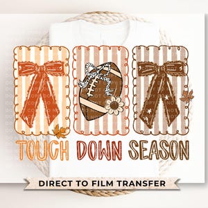 May include: A white t-shirt with a football-themed design. The design includes a football, bows, and the text "TOUCH DOWN SEASON". The design is in shades of brown, orange, and white, with a Direct to Film Transfer.