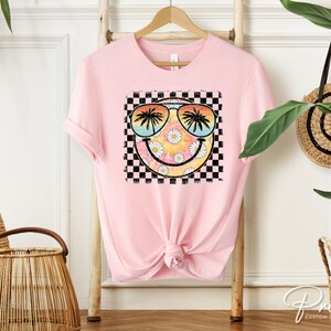 May include: Pink t-shirt with a graphic of a smiley face wearing sunglasses and palm trees. The smiley face is surrounded by a checkered pattern.