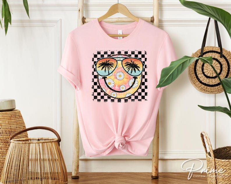 May include: Pink t-shirt with a graphic of a smiley face wearing sunglasses and palm trees. The smiley face is surrounded by a checkered pattern.
