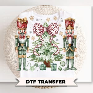 May include: White t-shirt featuring a Christmas design with two nutcrackers, a Christmas tree, and gifts. The design includes a large red and white polka dot bow, snowflakes, and the text "DTF TRANSFER" on a banner.