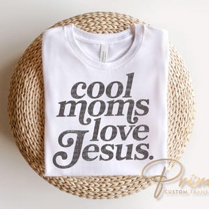 May include: White t-shirt with the text "cool moms love Jesus" in a dark gray, stylized font. The shirt is folded and rests on a woven, circular surface. The brand name "Prime Custom Transfers" is printed in gold.