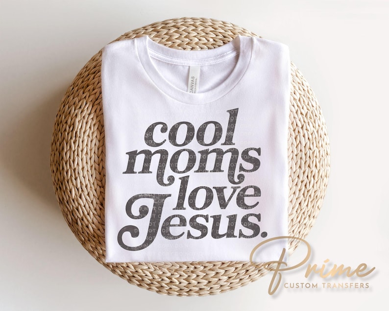 May include: White t-shirt with the text "cool moms love Jesus" in a dark gray, stylized font. The shirt is folded and rests on a woven, circular surface. The brand name "Prime Custom Transfers" is printed in gold.