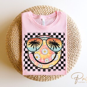 May include: Pink t-shirt with a black and white checkered background and a smiley face with sunglasses and palm trees. The smiley face is surrounded by daisies.