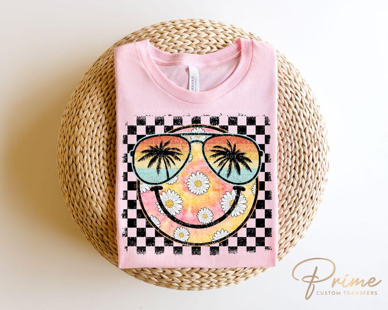 May include: Pink t-shirt with a black and white checkered background and a smiley face with sunglasses and palm trees. The smiley face is surrounded by daisies.