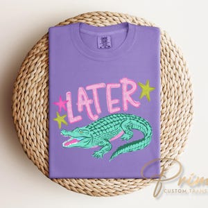 May include: A purple t-shirt with the word "LATER" in pink, outlined in white, and a green alligator illustration. Pink and yellow stars are on either side of the word. The shirt is folded on a woven, circular surface.