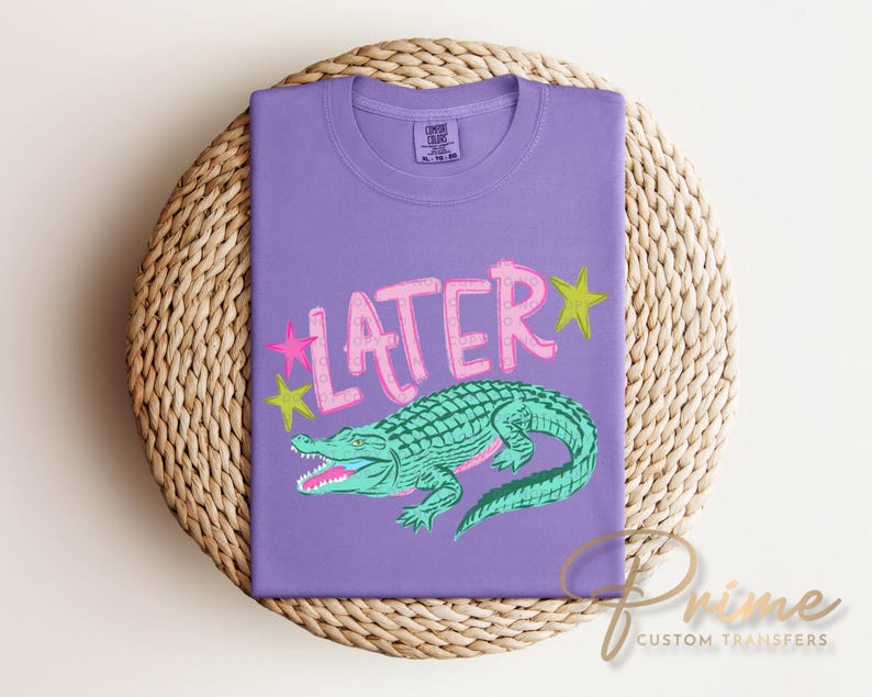 May include: A purple t-shirt with the word "LATER" in pink, outlined in white, and a green alligator illustration. Pink and yellow stars are on either side of the word. The shirt is folded on a woven, circular surface.