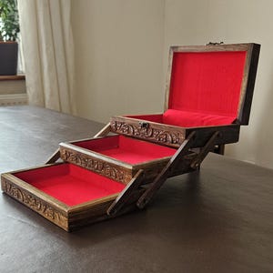 May include: A tiered wooden jewellery box with a dark brown finish and intricate carvings on the exterior edges. The box features three levels that expand outwards, each lined with a vibrant red fabric interior, including a hinged lid.