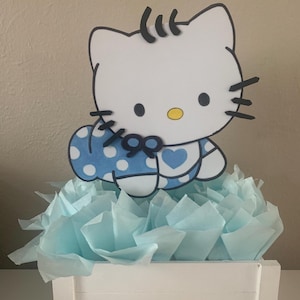 May include: A white Hello Kitty figurine wearing a blue and white polka dot dress with a blue heart on the front. The figurine is sitting on top of a white wooden box filled with light blue tissue paper.