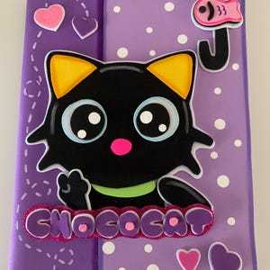 May include: A purple notebook with a cartoon black cat design. The cat has yellow ears, big blue eyes, and a pink nose. The word "CHOCOCAT" is written in glittery purple letters. White polka dots and heart shapes decorate the background.