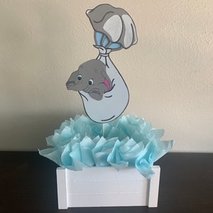 May include: A baby shower centerpiece featuring a white wooden box filled with light blue tissue paper. A cutout of Dumbo the elephant, in a blue and white balloon, is attached to a stick and placed in the center of the box.