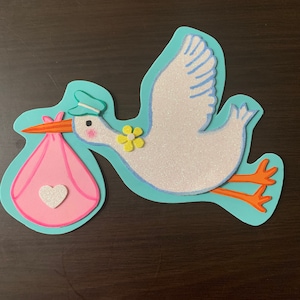 May include: A white stork with a blue hat and a yellow flower on its chest carries a pink baby bundle with a white heart on it. The stork is flying on a light blue background.