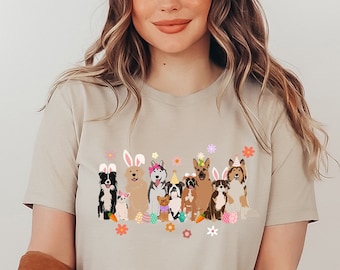 May include: A woman wearing a beige t-shirt with a graphic of dogs wearing bunny ears and holding Easter eggs. The dogs are all different breeds and colors. The graphic is surrounded by flowers.