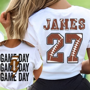 May include: White t-shirt with brown football design and the text "JAMES 27" on the back. The front of the shirt has the text "GAME DAY" repeated three times with a lightning bolt and football design.