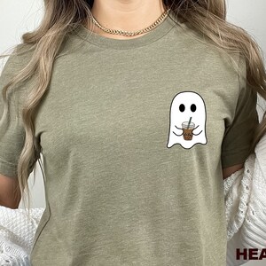 Puede incluir: Una camiseta de color oliva jaspeado con un gr&aacute;fico de un fantasma sosteniendo una taza de caf&eacute;. El texto "HEATHER OLIVE" est&aacute; impreso en la esquina inferior derecha de la imagen.