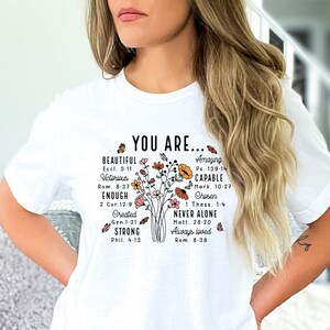 May include: White t-shirt with a floral design and the text "YOU ARE..." surrounded by words and bible verses, including "Beautiful", "Victorious", "Enough", "Created", "Strong", "Amazing", "Capable", "Chosen", "Never Alone", "Always Loved".