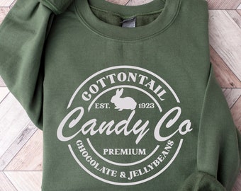Könnte beinhalten: Ein olivgrüner Sweatshirt mit einem weißen Kreislogo, auf dem "Cottontail Candy Co. Est. 1923 Premium Chocolate & Jellybeans" steht. Das Logo zeigt auch eine weiße Silhouette eines Kaninchens.
